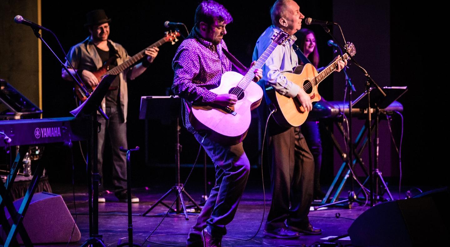 A side-on image of Al Stewart on stage with a guitar, peforming with other musicians around him