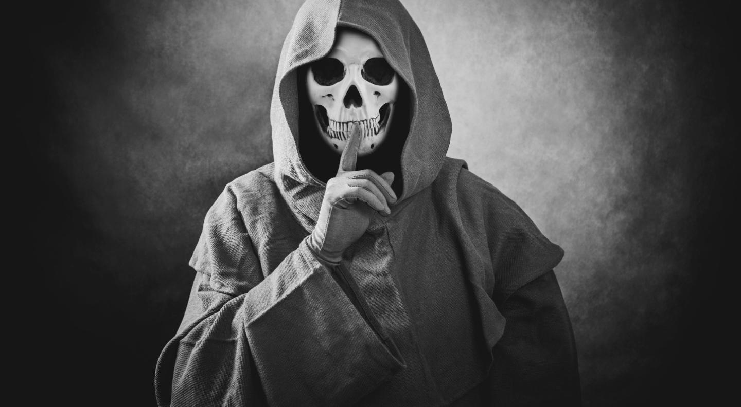 A black and white image of a skeleton wearing a grey robe. He holds his finger to his lips