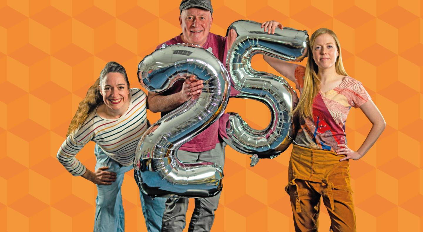 Three people standing against a bright orange background holding silver balloons in the shape of the number 25