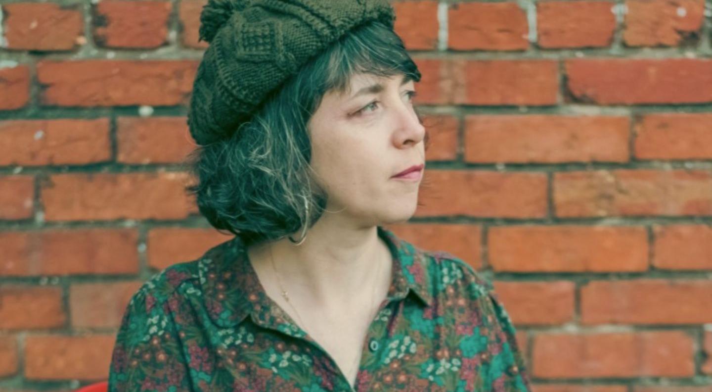 Lisa O'Neill wears a floral shirt and a knitted woolen hat. She looks off to her left against a red brick background