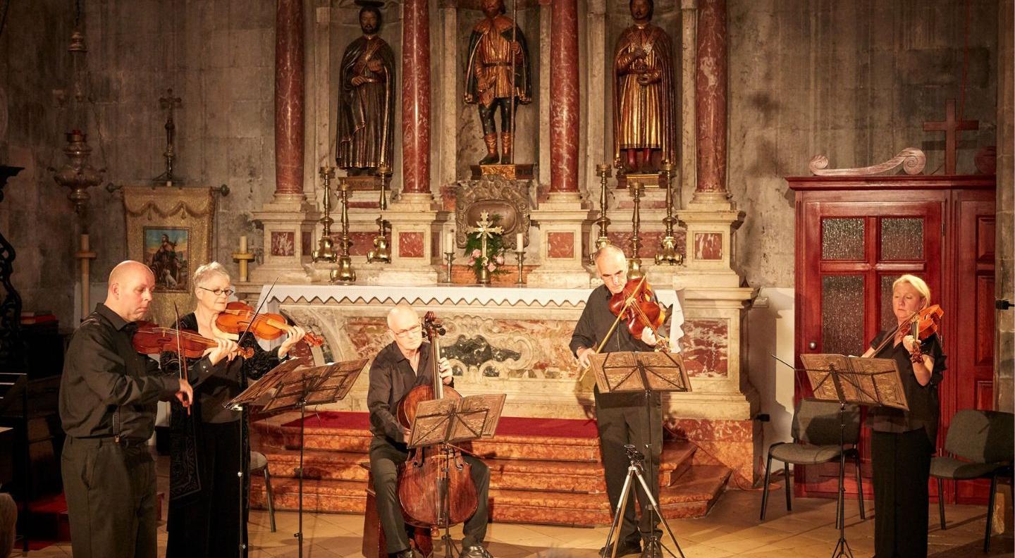 A group of musicians play string instruments in a very ornate and old catholic church, surrounded by religious statues and mosaics