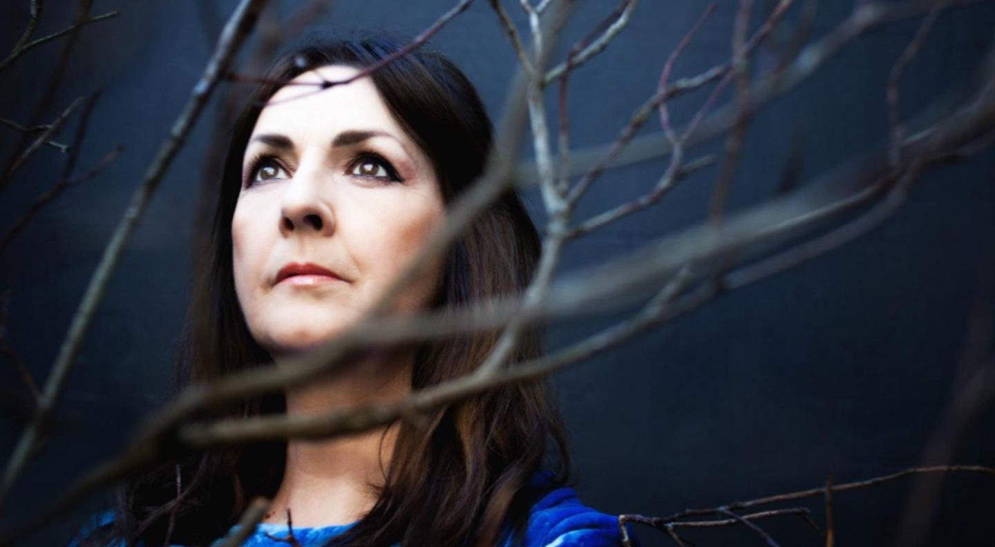 A white woman with dark hair and a blue shirt stands among the bare branches of a tree, looking upwards