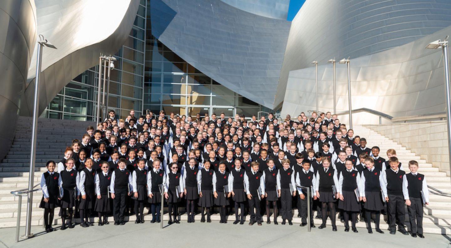 A large group of children wearing NCC uniforms stand in front of the Walt Disney Concert Hall in LA, a large silver building