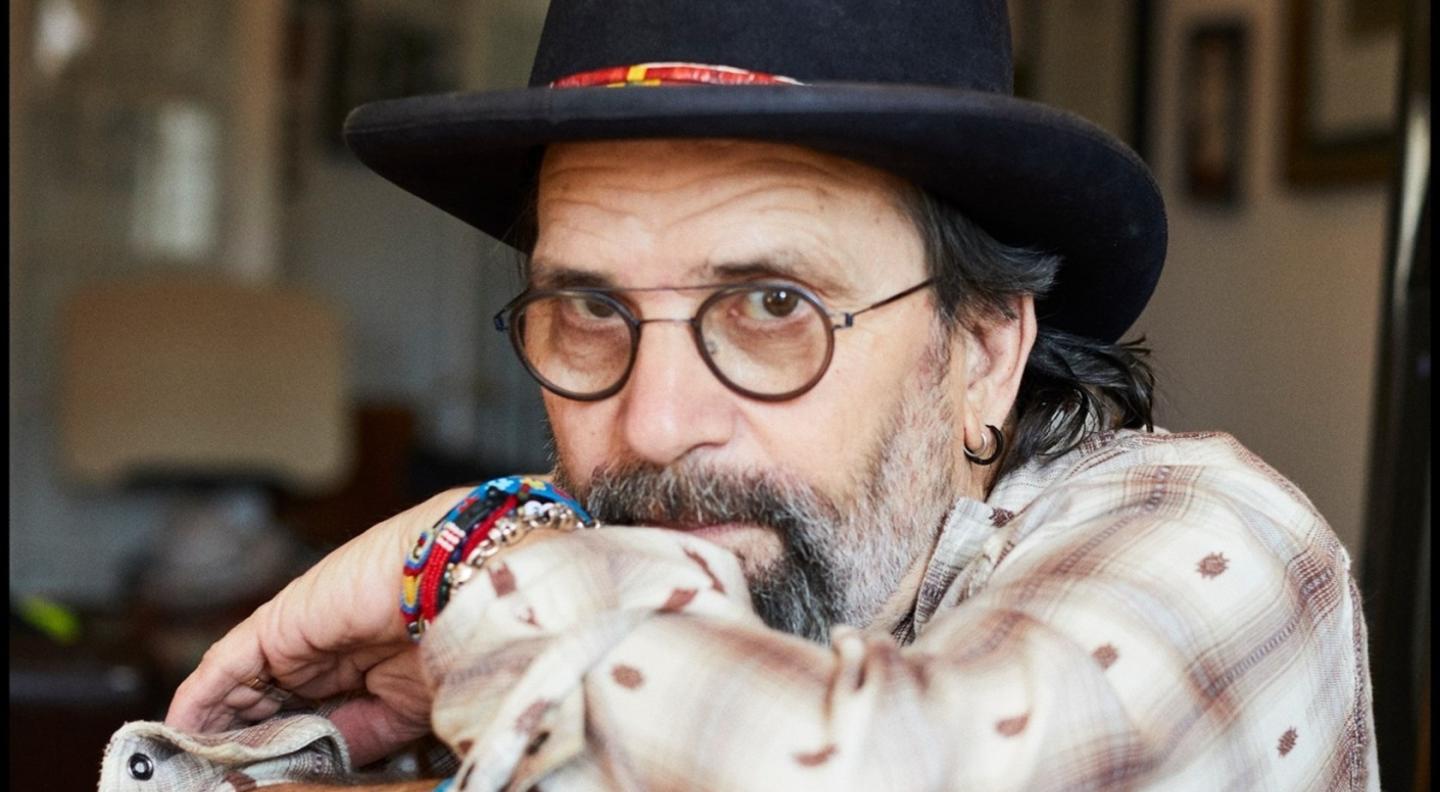 A close up image of Steve Earle wearing a hat and staring into the camera over his hand
