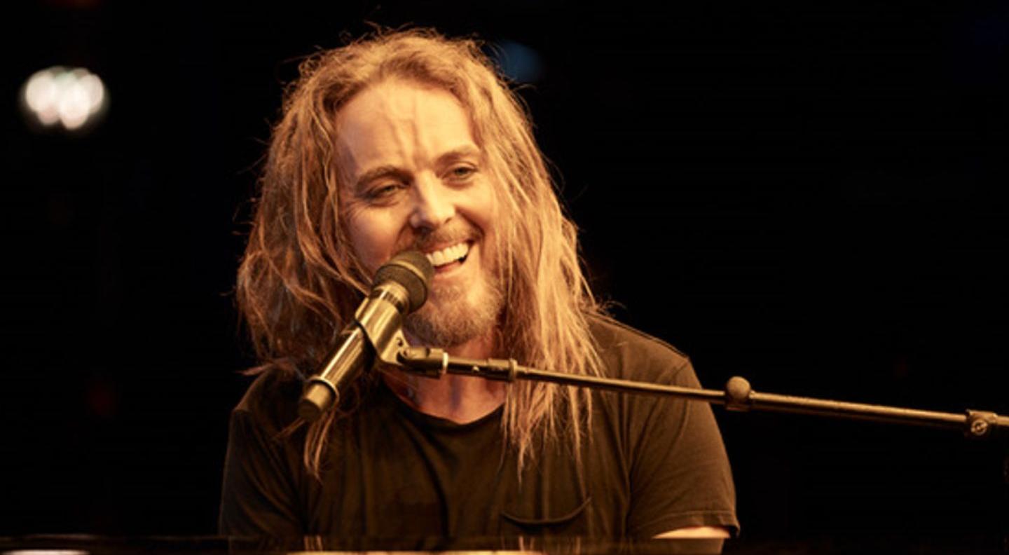 A white man with long ginger hair and a beard sits, smiling widely, at a piano with a microphone