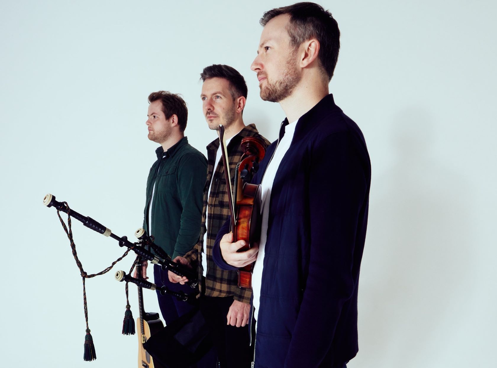 Three young white men stand in a diagonal line holding a fiddle, pipes and guitar