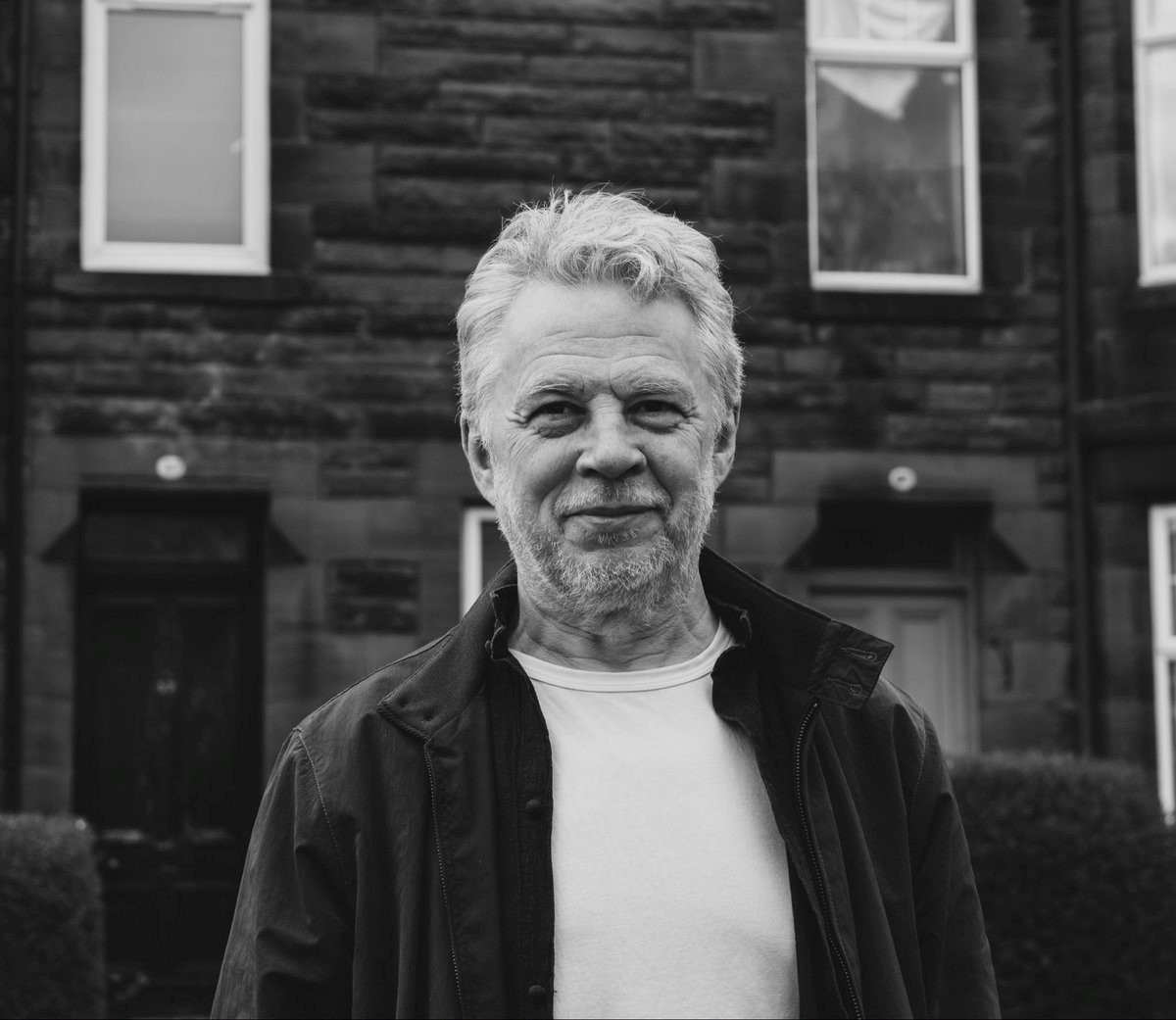 A black and white photo of a white man with silver hair standing in front of a brick building with windows