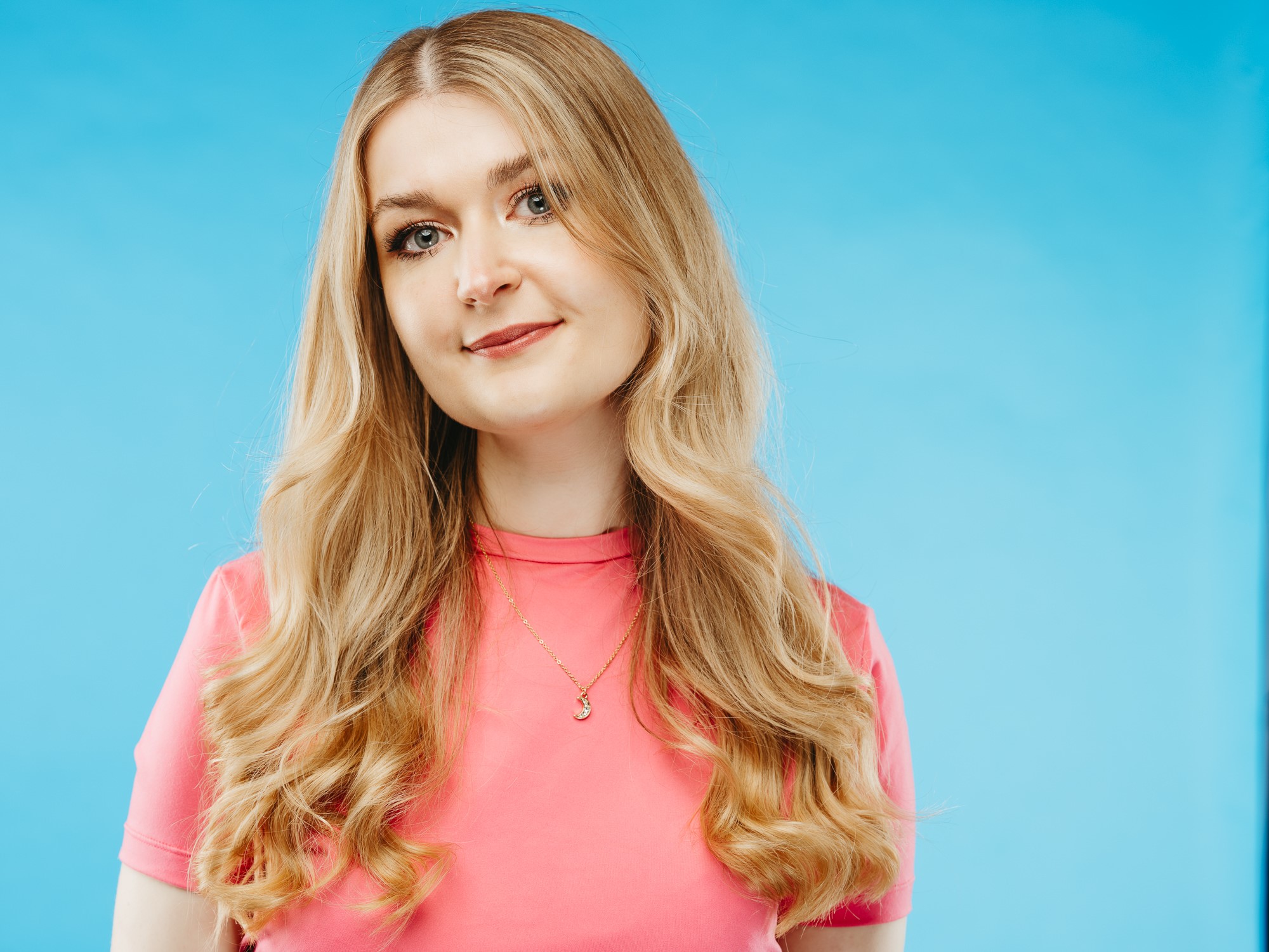 A young white woman with long blonde wavy hair wearing a pink top standing in front of a blue background