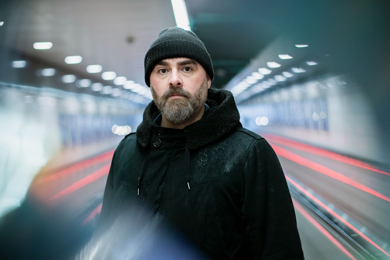 A white man with a dark beard and wearing dark clothes stands looking at the viewer as light refracts around him