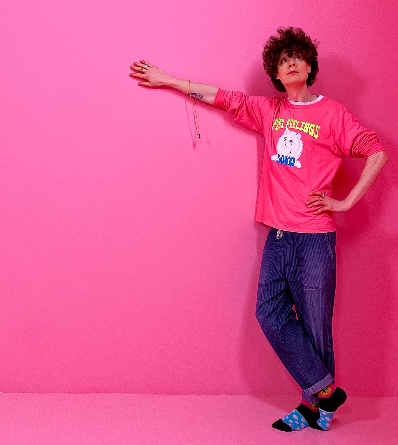 Michael Pedersen, a white man with curly dark hair, stands against a bright pink background with one arm leaning on the back and one on his hip. He wears dark trousers and a bright pint sweatshirt with a white image