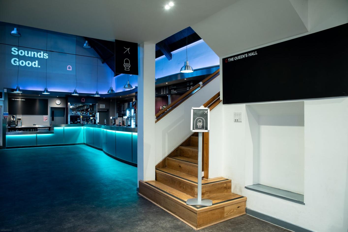 A large bar room with a staircase, coloured lights and shelves of drinks