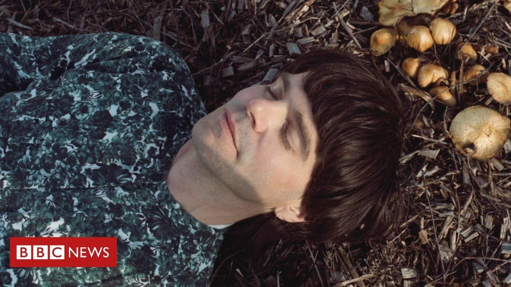 A man with dark hair and a flowery shirt lies on his back with his eyes closed. There are mushrooms growing out of the earth above his head