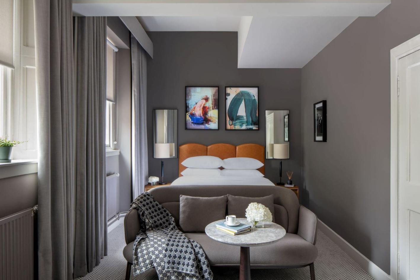 A luxurious hotel bedroom with grey decor, a large bed, colourful paintings, and assorted furrniture, soft furnishings and lamps