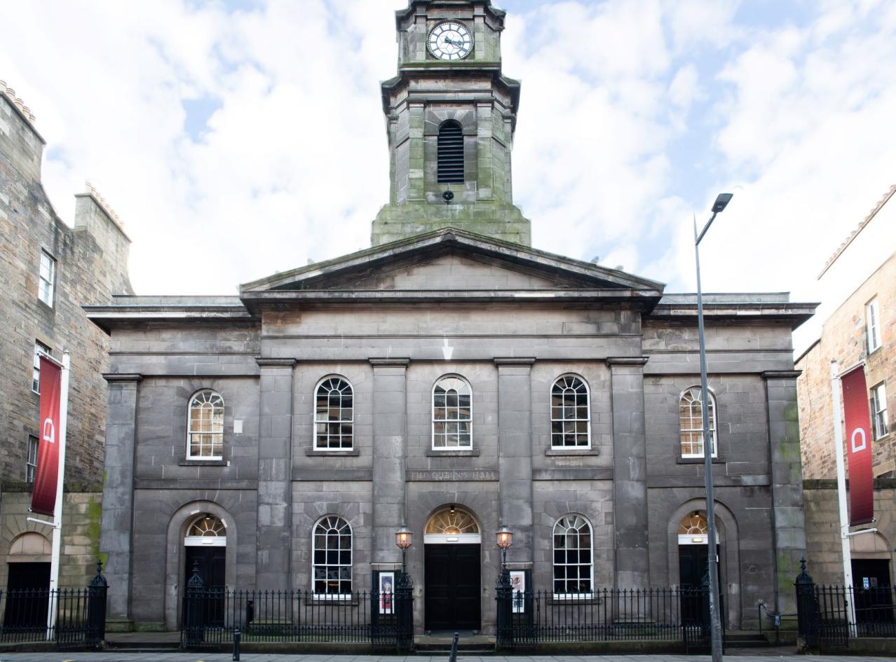 The outside of The Queen's Hall a large grey stone building with red flags at either end, a tower with a clock in the middle and lots of doors and windows