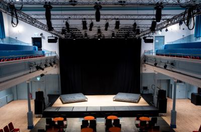 A black backdrop over a stage with two raised plaforms and tables and chairs in front. Also shows a balcony area and lighting rig