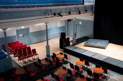 Side view of a stage with a black backdrop and a raised plaform with tables and chairs in front. Also shows a balcony area and lighting rig