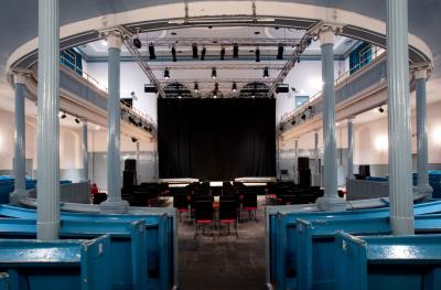 Interior of a modern theater with blue seating, a black stage, and professional lighting rigs, set in a historical building with white columns and an ornate ceiling.