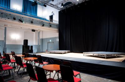 A black backdrop hangs over a stage with two raised plaforms. Tables and chairs are in front of the stage