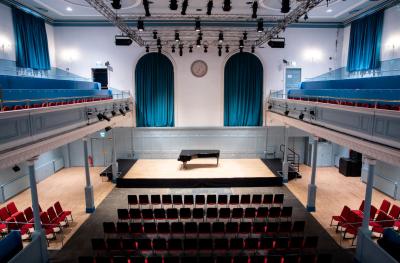 A view from above of a stage with a grand piano on it and a black backdrop ,rows of red seats in front, seats on a gallery and a lighting rig