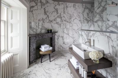 A luxurious hotel en-suite bathroom with white marbled walls and floor, white hand basin and towels and bottles of bath products