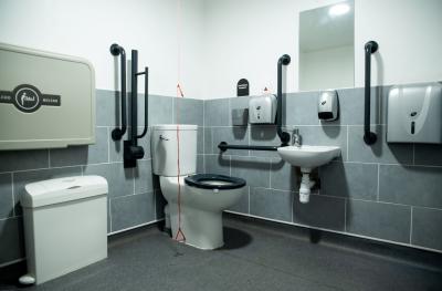 The inside of an accessible toilet with grab bars, a baby change table, an emergency cord and sink and hand towels