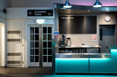 A green lit bar with a coffee machine behind it and double doors to the side with a sign above that reads Accessible Toilets with an arrow pointing left