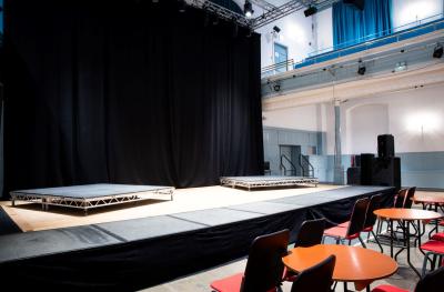 An empty stage with a black backcloth and red chairs around round tables in front