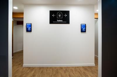 A white wall with a black sign showing a toilet icon and arrows pointing to the right and left. Text reads Toilets. Please use any cubicle then exit at the other end.