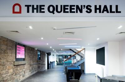 A red arch and the words The Queen's Hall are written in bold text on a wall with a view down a long foyer underneath with stone walls, grey flooring, bright lights and a TV screen