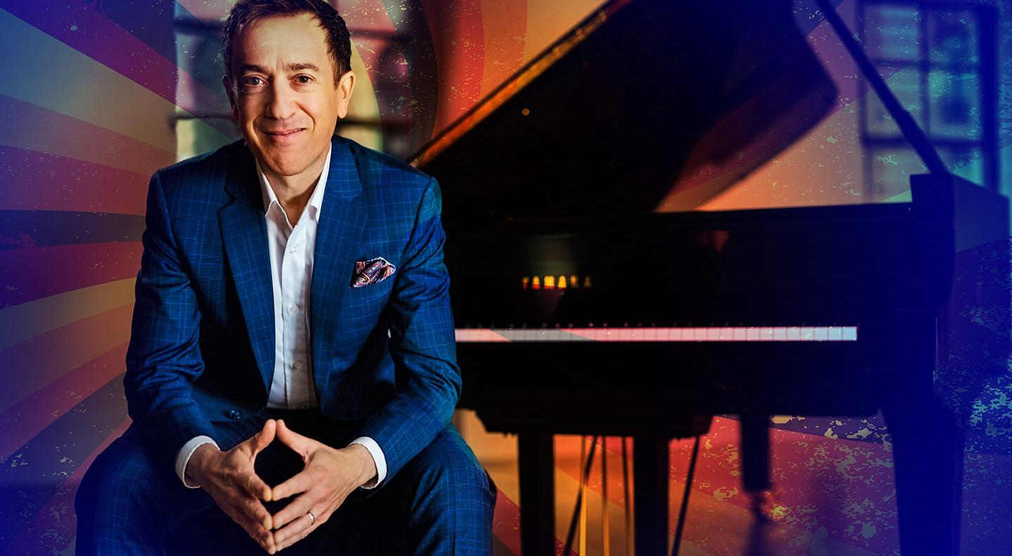 Geoffrey Keezer, a white man with dark hair sits by a grand piano with his arms resting on his knees, smiling at the camera