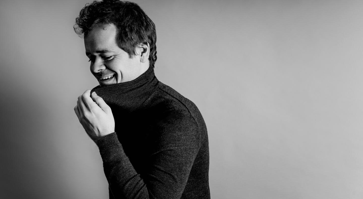 A black and white mid shot of Benjamin Grosvenor pulling at the neck of his black rollneck jumper and laughing into it with his eyes closed