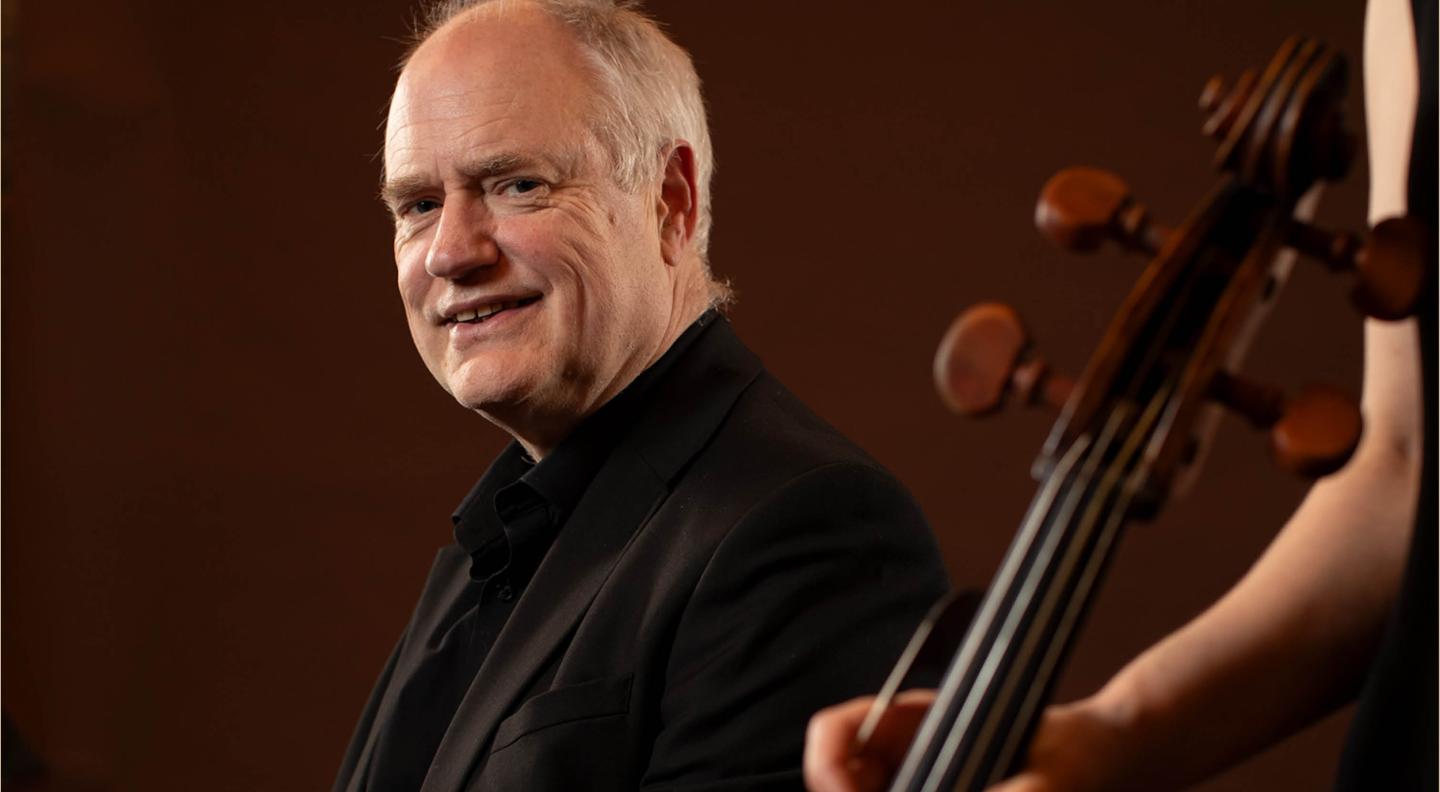 a bald man looks toward the camera against a dark red background. A hand holding the neck of a violin is in the foreground
