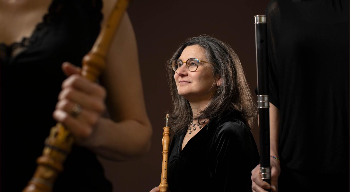 A white woman with shoulder length greying hair and glasses sits in the centre of the image holding a wind instrument. To either side of her in the foreground are the torso and arms of other people holding wind instruments