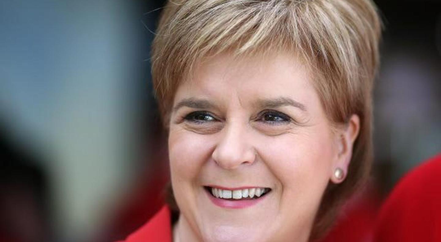 A close up shot of Nicola Sturgeon, a white woman with short fair hair. She is smiling and wears a bright red top