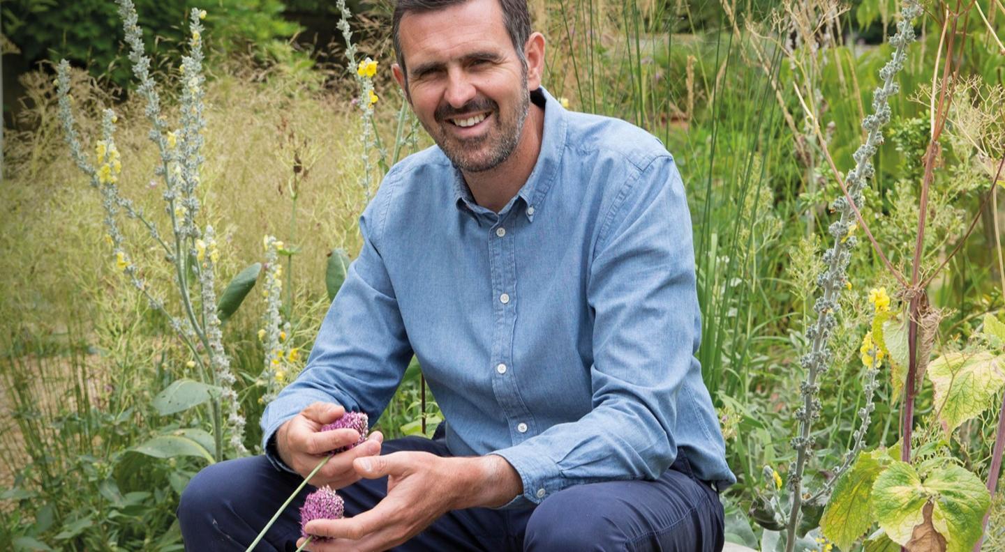 Adam Frost sits in a garden, holding a flower in his hand