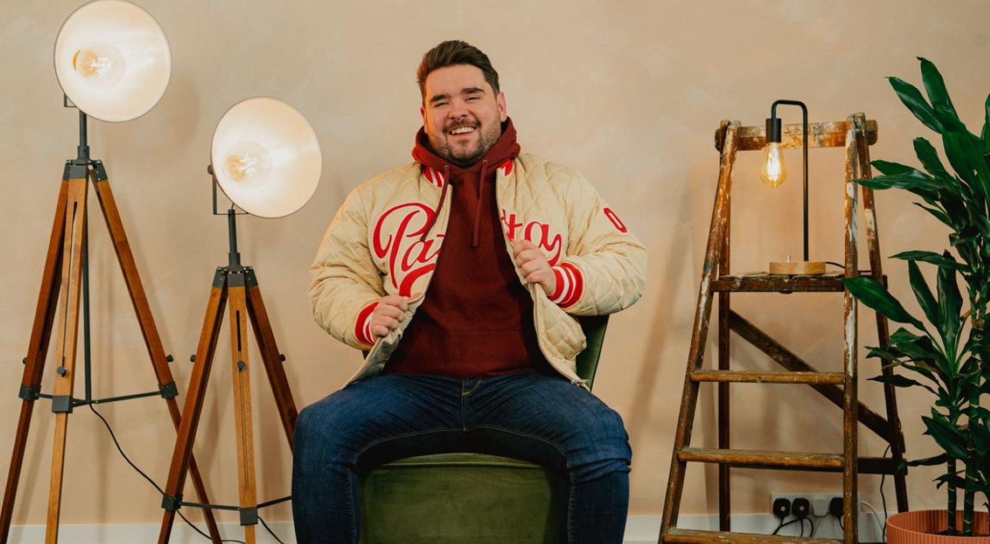 Adam Rowe, a young man with dark hair and a beard sits on a green velvet chair smiling. There are two bright floor lights on his right and a stepladder and a plant on his left