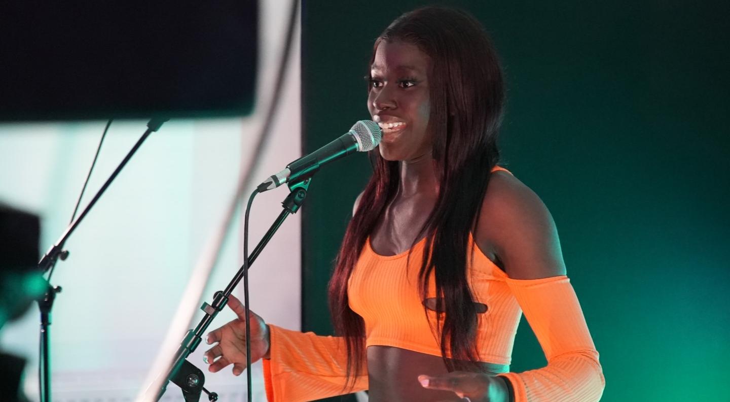 AiiTee, a young black musician, sings at a microphone. She wears a bright orange jumpsuit