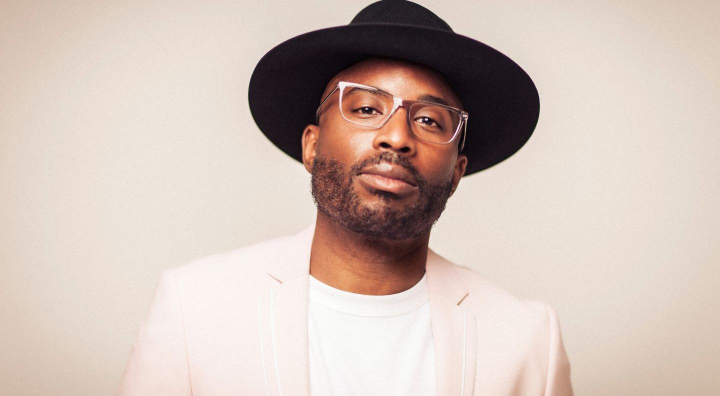 Alexis Ffrench wears a cream suit and white -shirt and black wide-brimmed hat and glasses, and stands against a cream background