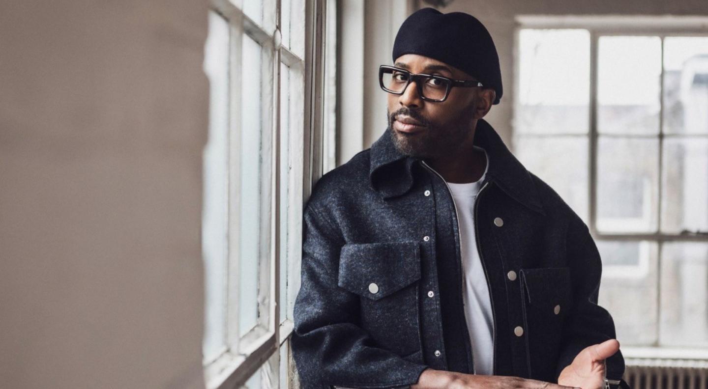 Alexis Ffrench, wearing a black leather jacket over a white t-shirt and a black kangol hat, looking at the camera in front of a window