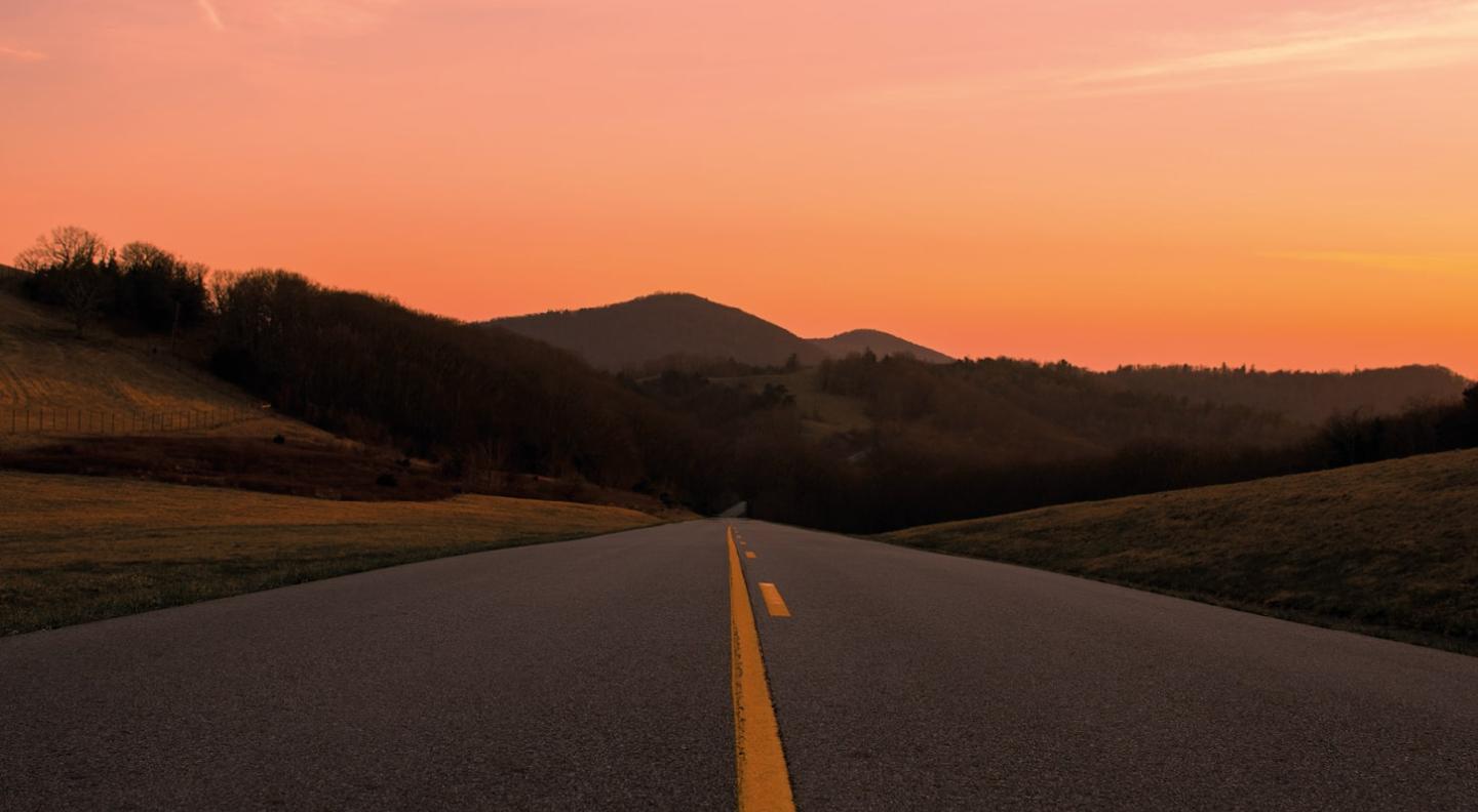 A view of a wide open road with a yellow line down the centre which disappears off into trees in the distance surrounded by an orange sky