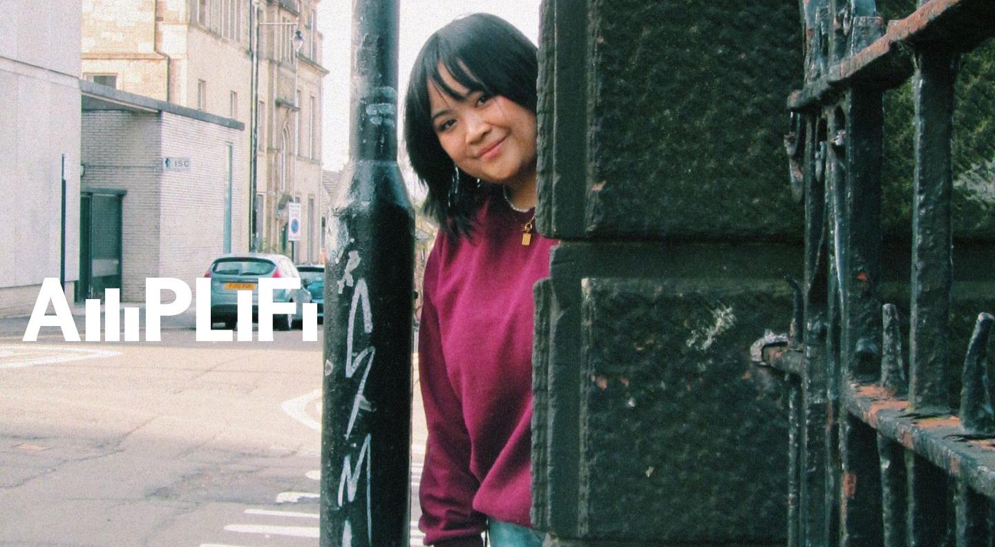 Belly Rachel, a young Malaysian woman with short dark hair, peers out from behind a wall. Text reads 'AMPLIFI'