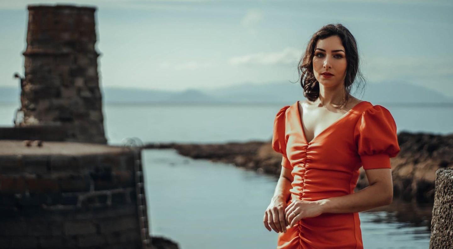 am image of Chloe Matharu, a young Scottish-Indian woman, against a backdrop of a harbour. She wears a bright orange dress