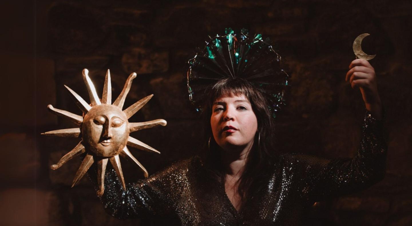 Bell Lungs (Ceylan Hay) holding a model of the sun in one hand, and a moon in the other, standing in front of a brick background