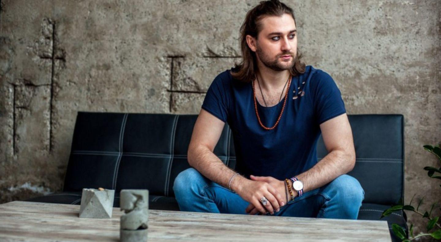 A young white man with long dark hair and stubble sits on a sofa against a concrete background. He has heavy eyeliner and wears a dark t-shirt and jeans.