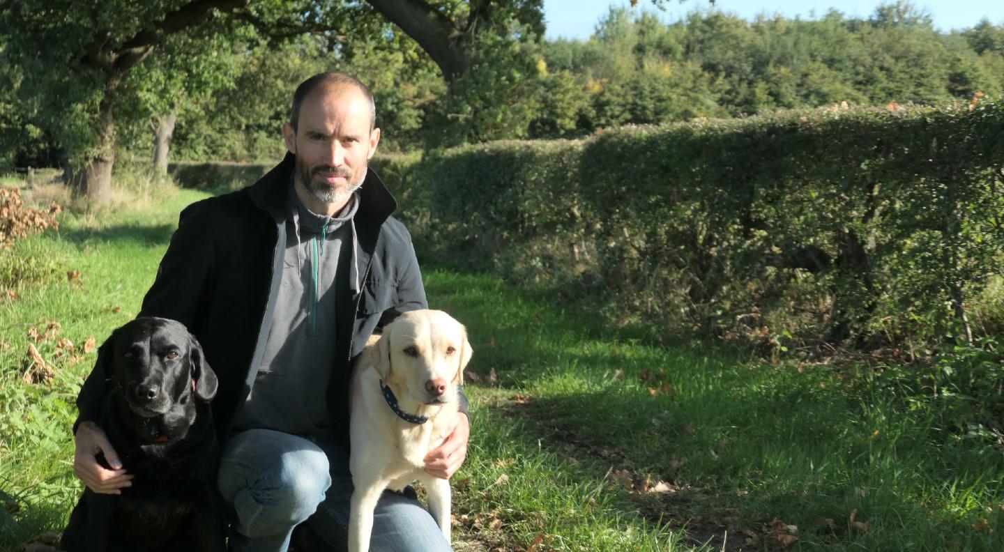 Andrew Cotter kneels down in a field with his dogs Oliver, a black labrador, and Mabel, a golden labrador