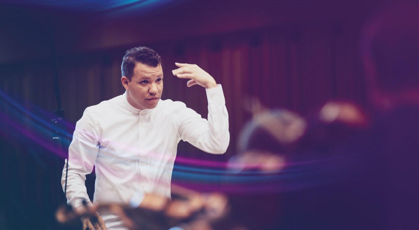 A young white man with short brown hair conducts an orchestra, wearing a white shirt