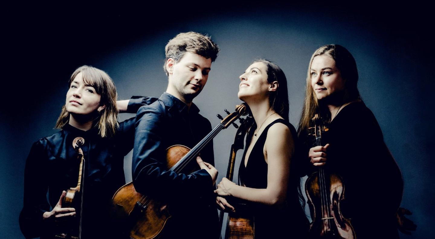 The Barbican Quartet - three white women and one white man - stand wearing dark clothing holding their string instruments in front of a dark background, gently surrounded by a halo of light