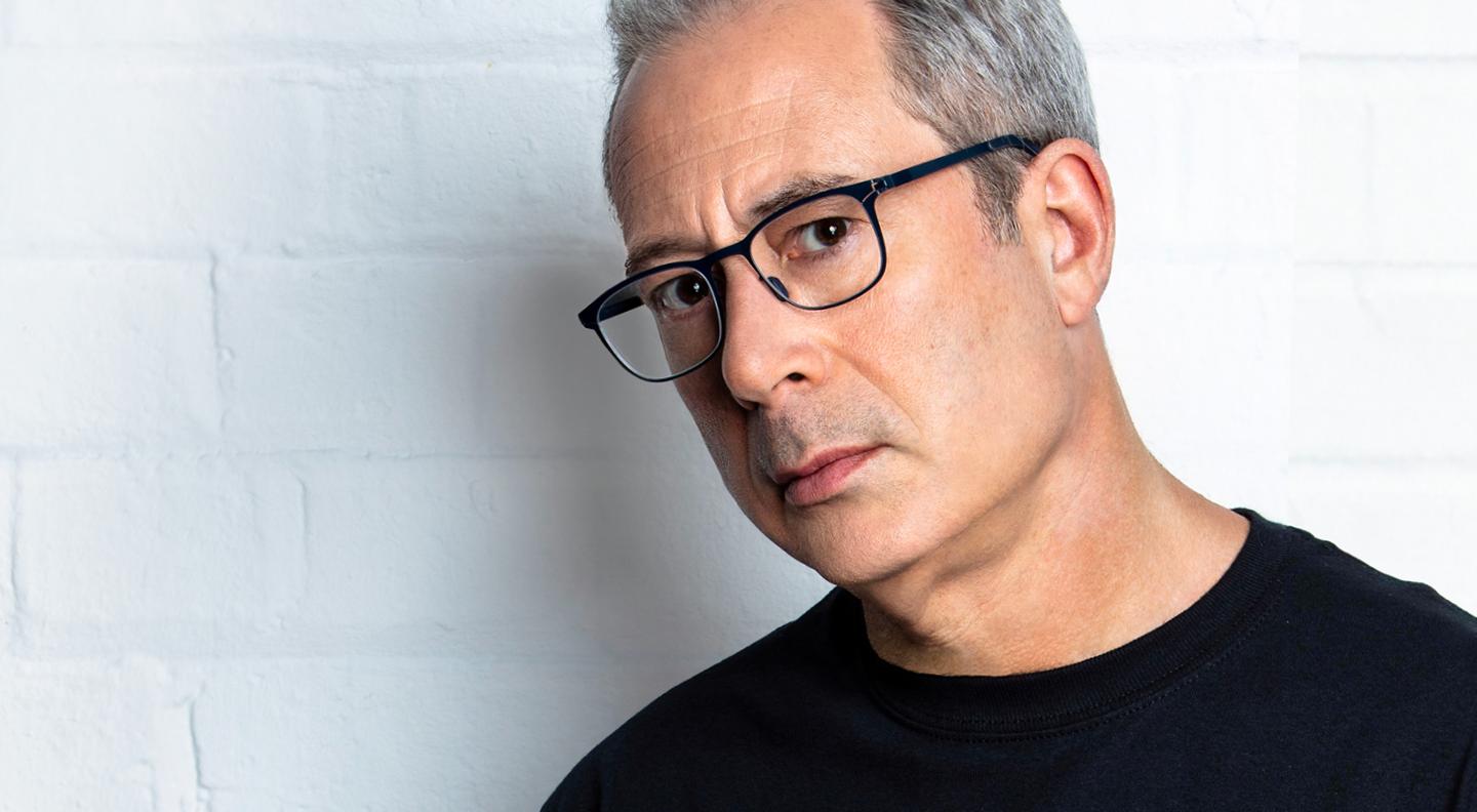 An image of Ben Elton, a white man with grey hair. He wears a black t-shirt and black rimmed glasses