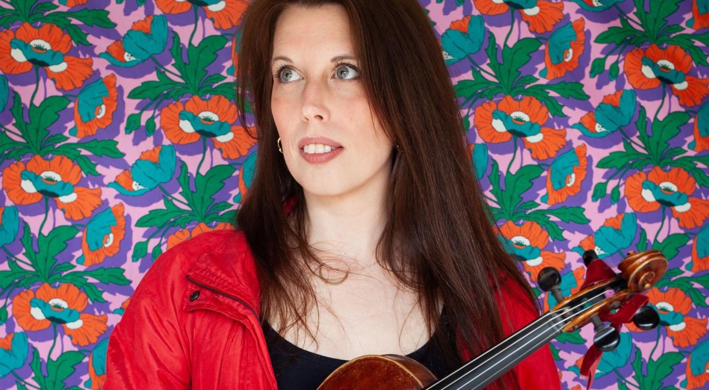 A white woman with long dark hair wearing a red jacket and holding a fiddle stands in front of a flowery patterned background in red, purple and green