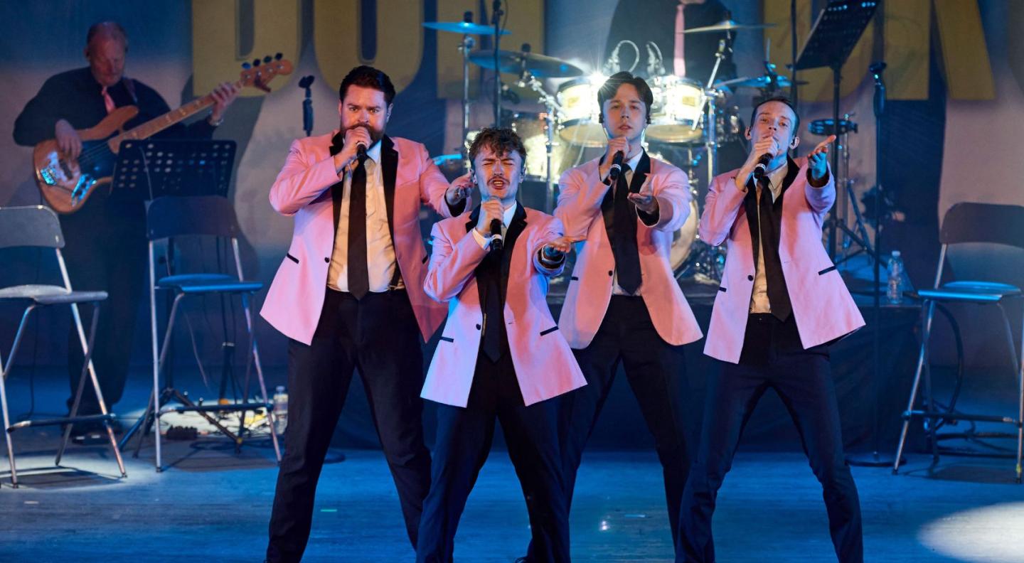 The four members of Big Girls Don't Cry performing on stage. All members are male, and are wearing suits with pink blazers.